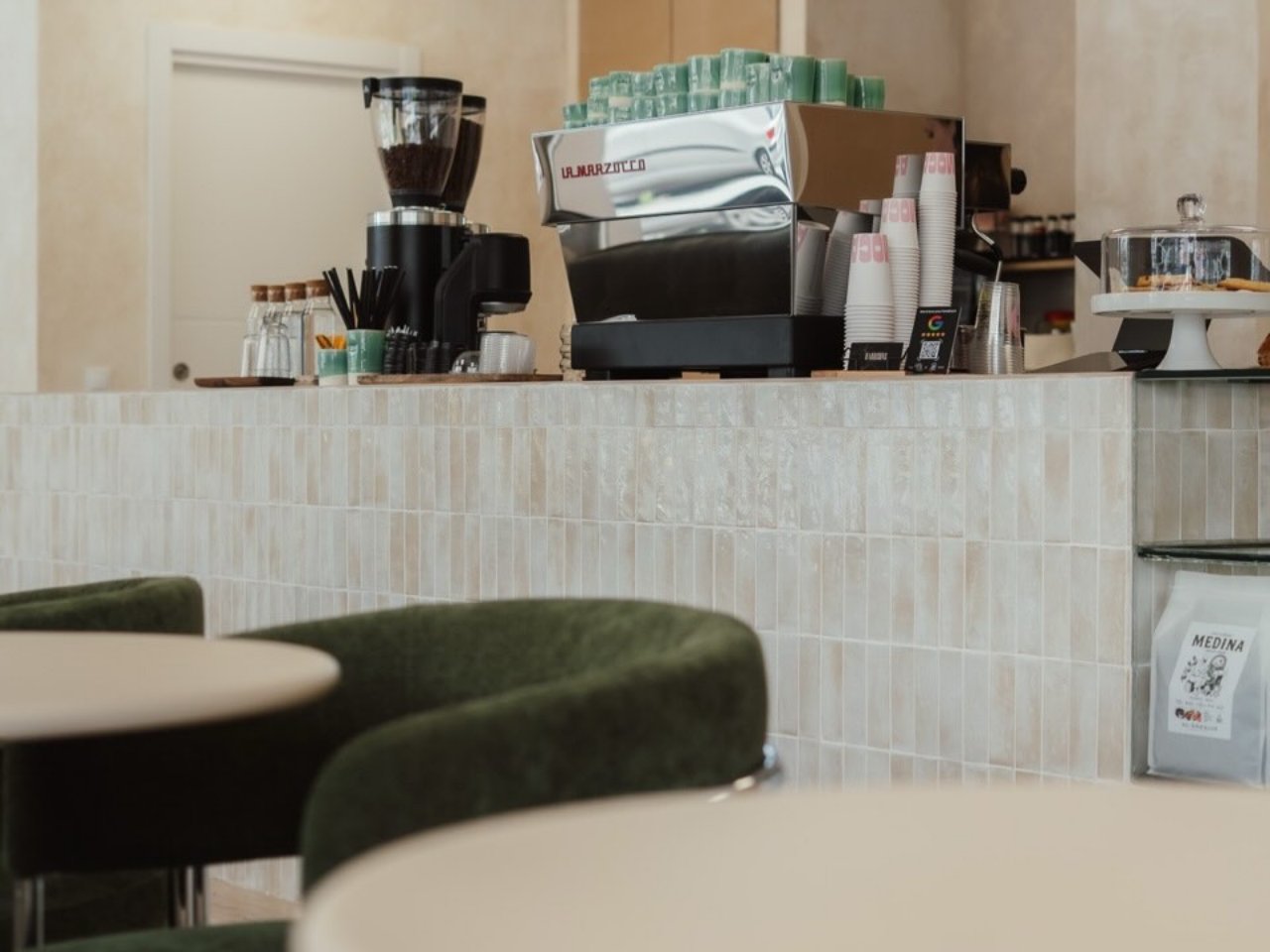 La barra de Moca con la maquina La Marzocco y el molinillo, con las mesas y sillas de terciopelo verde delante